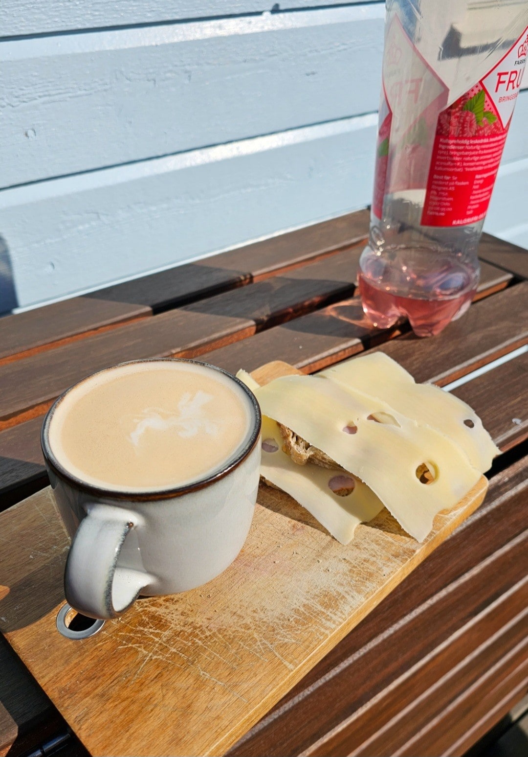 A small ceramic cup contains a light-colored beverage, placed on a wooden cutting board. Nearby, slices of cheese rest next to the cup. A plastic bottle of a pink liquid is visible in the background, complementing the outdoor setting.