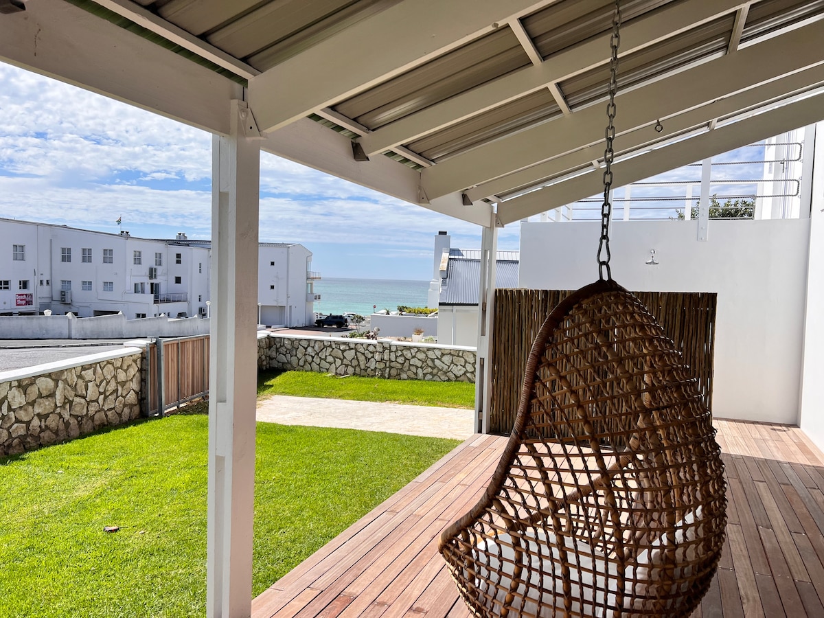 A woven hanging chair is suspended from a shaded patio, overlooking a grassy area that leads to a picturesque beachfront view. A pathway of light-colored stones and a stone wall can be seen in the background, with calm waters visible in the distance.