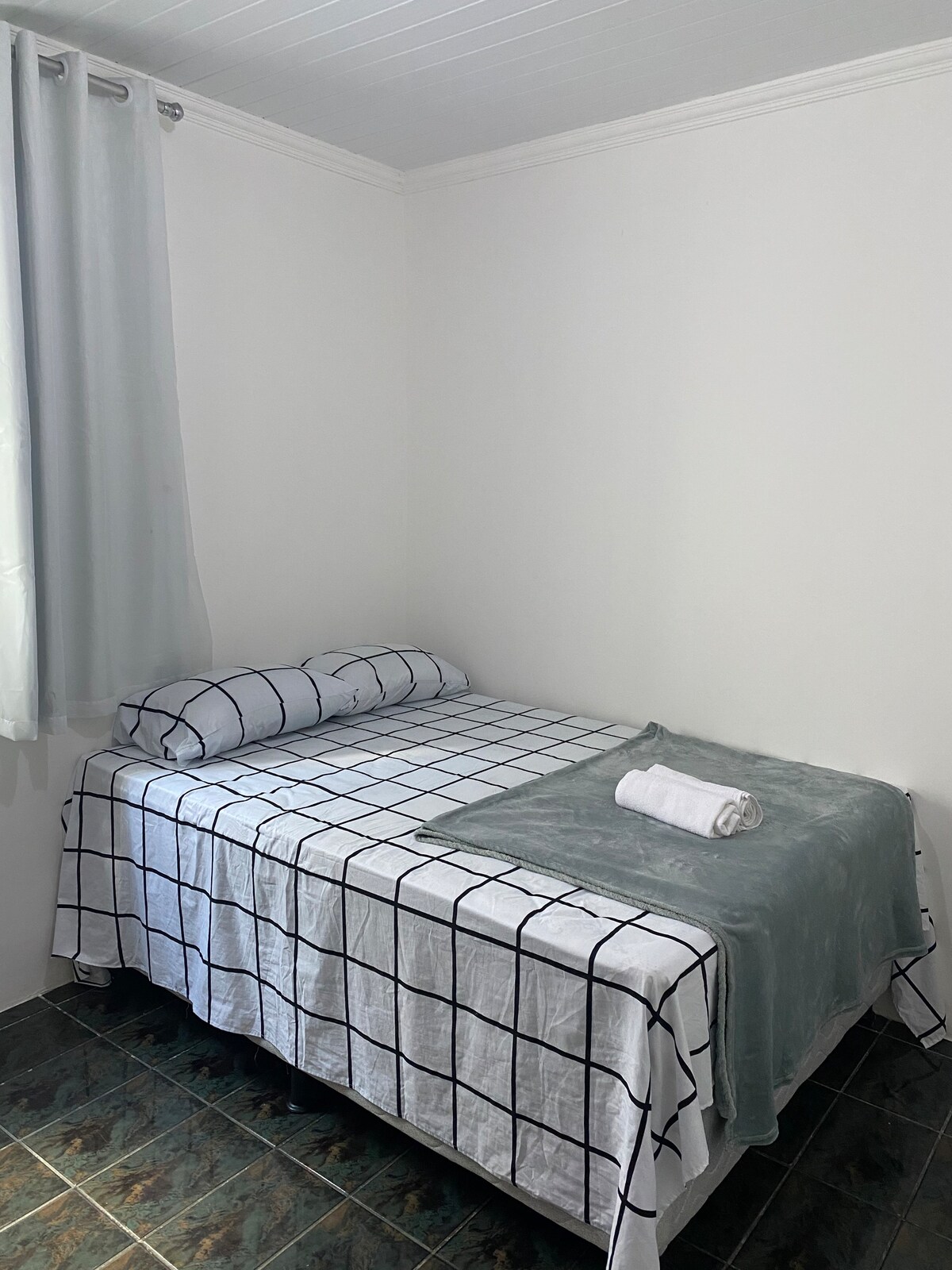 A well-arranged sleeping area features a double bed dressed in a grid-patterned coverlet. A soft throw blanket and a rolled towel are placed neatly at the foot of the bed. Light filters through a sheer curtain, adding a gentle brightness to the space.
