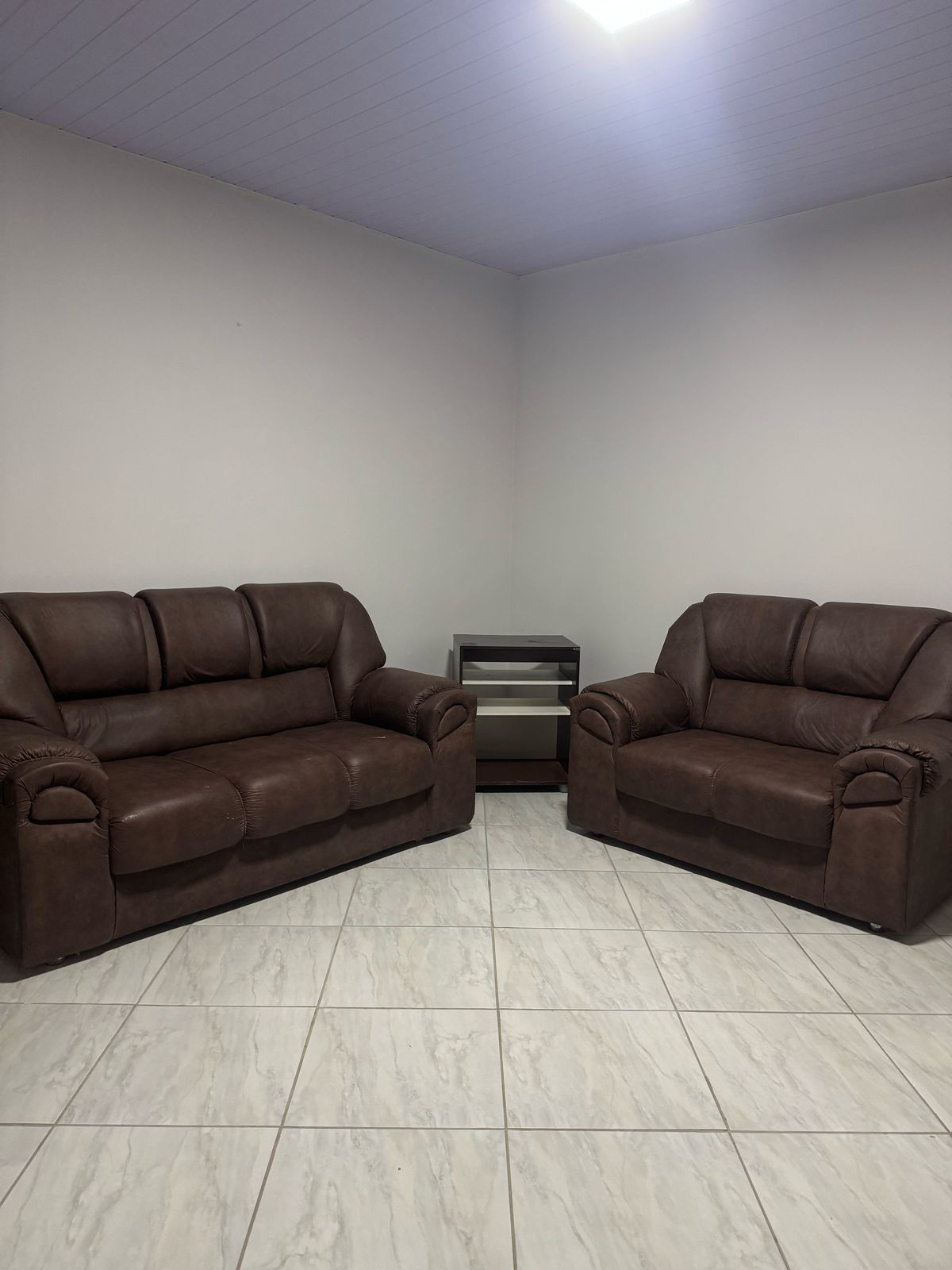 A cozy seating area features a pair of brown sofas positioned in the corner of the room. A small, dark side table is visible between the sofas, complementing the neutral tile flooring and light-colored walls.