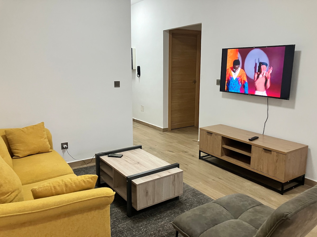 The living room features cozy seating, including a yellow three-seat sofa and a contrasting gray area rug. A wooden coffee table is centrally placed. A wall-mounted smart TV displays a vibrant image, while the entrance to the hallway is visible in the background.