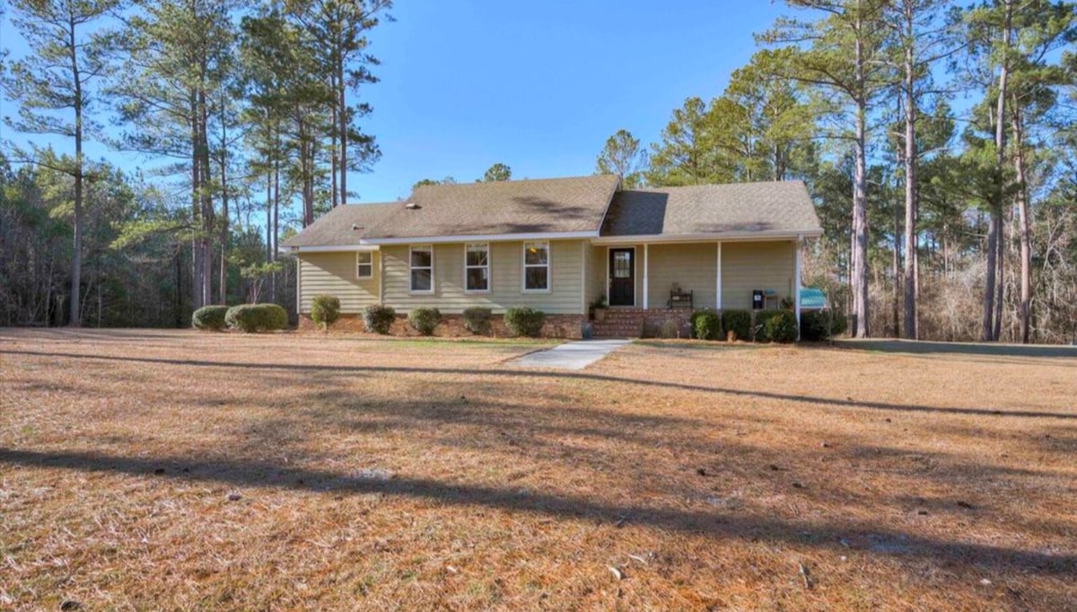 A single-story home is set amidst tall trees and open grassland. The exterior features large windows and a welcoming front porch. A well-maintained lawn is visible, framed by neat shrubs and a long driveway leading to the front door.