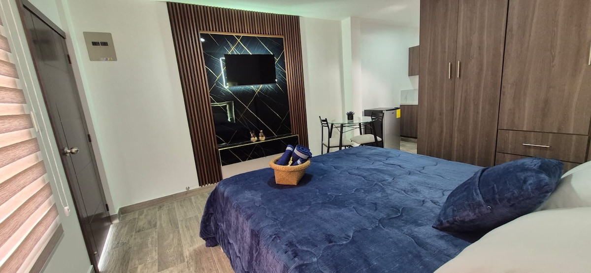 A spacious sleeping area features a luxurious bed with a rich blue cover and decorative pillows. A wall-mounted TV is set against a stylish backdrop with geometric patterns. A modern dining table and chairs are positioned nearby, complementing the elegant design of the suite.