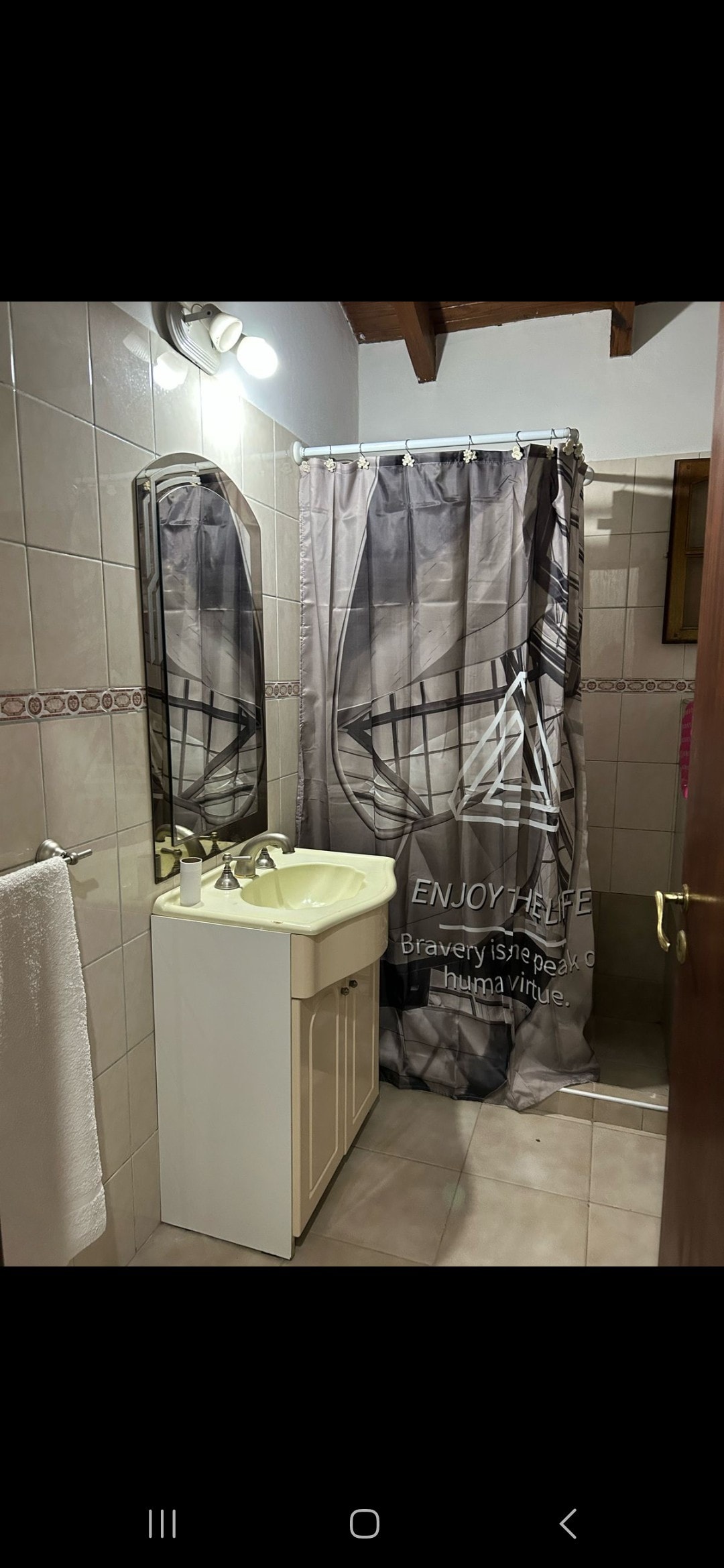 The bathroom features tiled walls and a shower area separated by a patterned curtain. A small sink with cabinet storage is visible, along with a towel hanging from a rack. Soft lighting is provided by overhead fixtures.