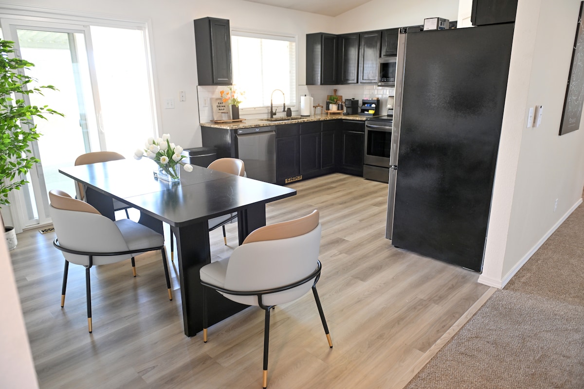 A modern kitchen and dining area features a sleek black dining table with four upholstered chairs. Light wood flooring extends throughout, complemented by black cabinetry and stainless-steel appliances. Natural light enters through sliding glass doors, creating a welcoming atmosphere.