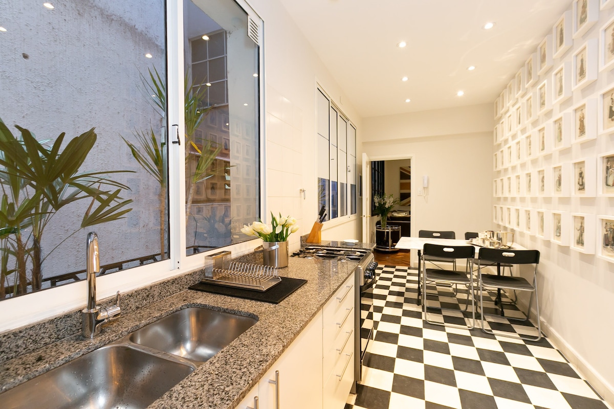 A modern kitchen features a spacious granite countertop with a double sink and sleek appliances. Bright, natural light floods through large windows adorned with a view of greenery. A dining area is positioned alongside a wall decorated with framed photos, complemented by a table and black chairs.