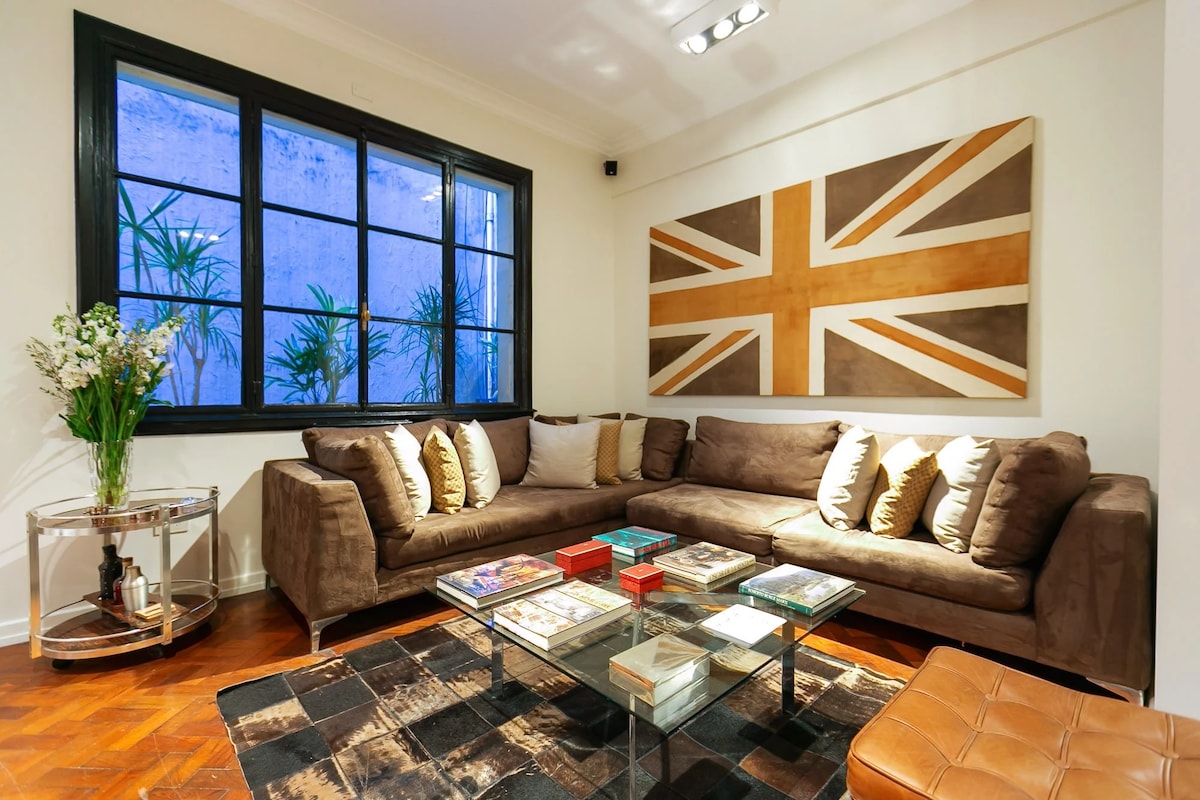 A spacious living area is captured, featuring a large sectional couch adorned with decorative cushions. A glass coffee table holds assorted books and magazines, while a round side table displays a floral arrangement. Natural light filters through a large window overlooking greenery.