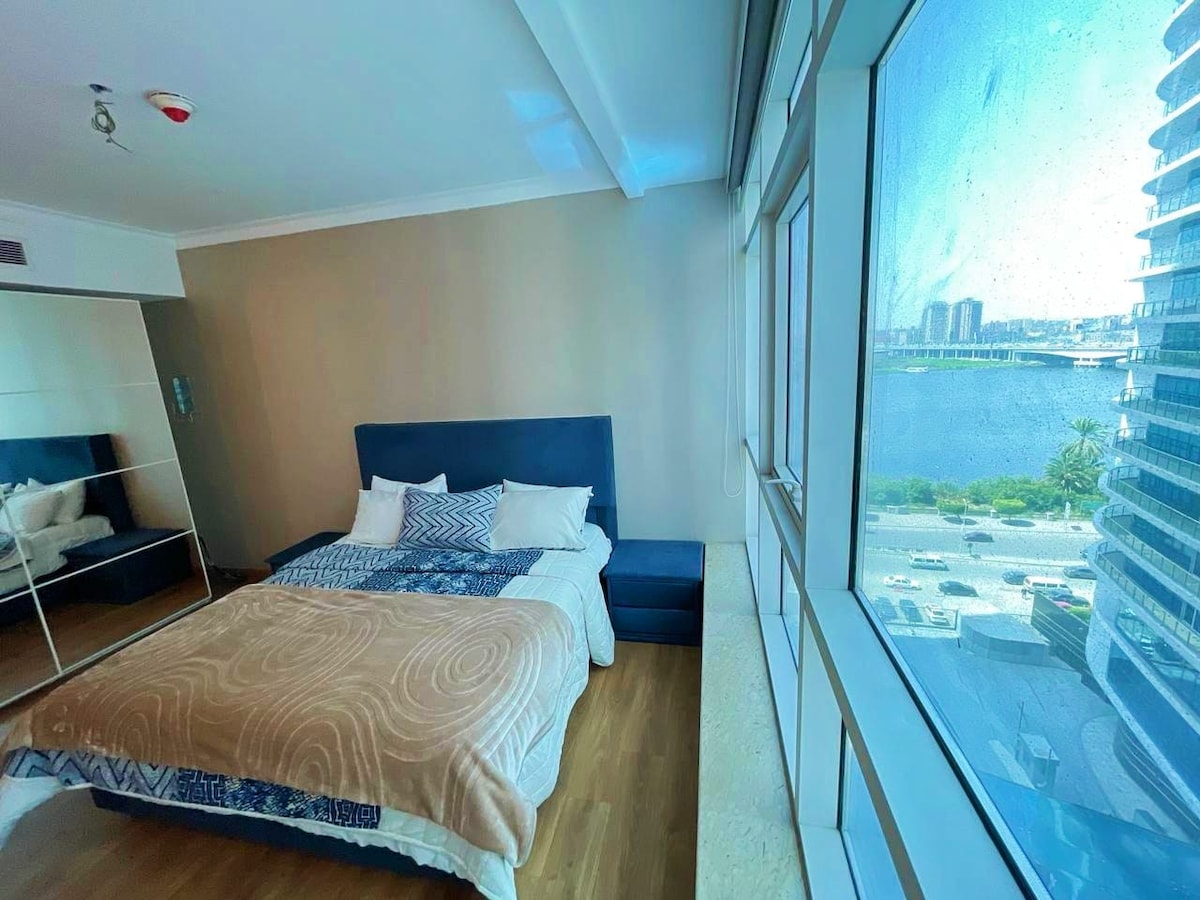 A spacious bedroom features large windows that offer views of the water and skyline. A generous bed is complemented by clean lines and a neutral color palette. A seating area is positioned near the window, enhancing natural light within the room.