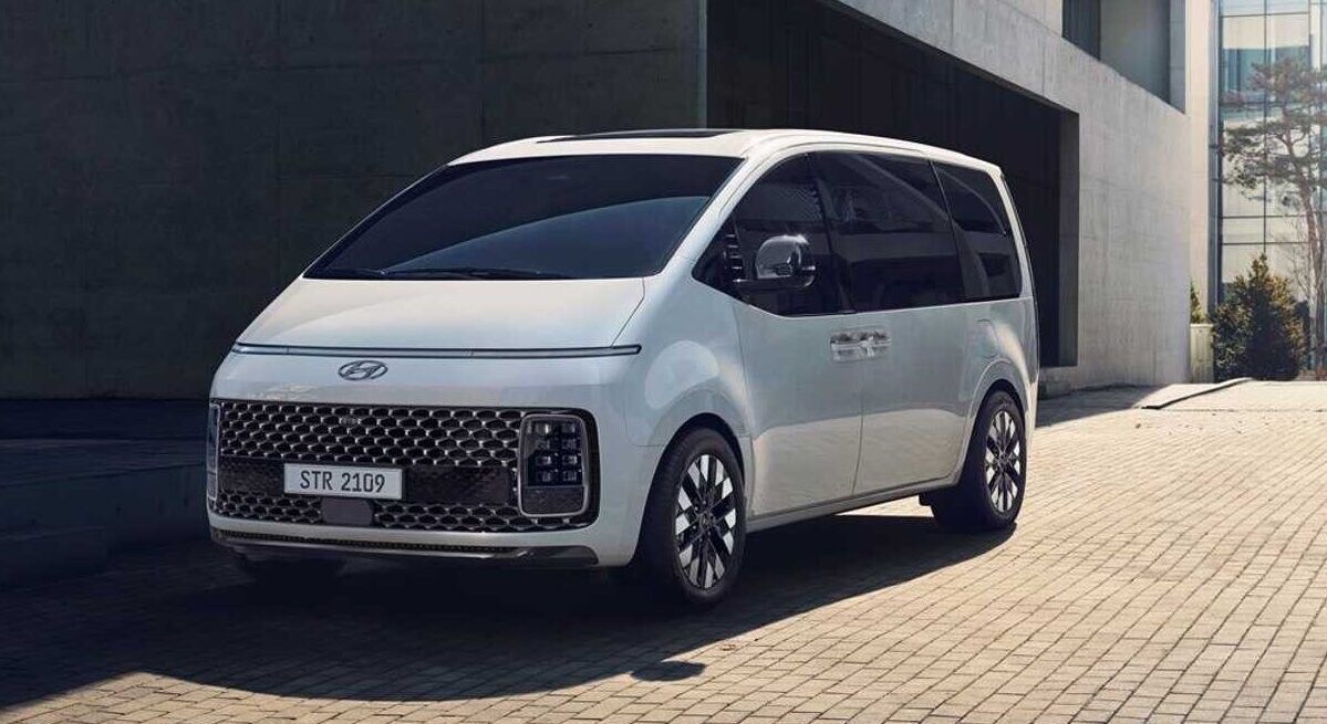 A modern white van is parked on a textured stone surface, showcasing a sleek and angular design. Its front features a distinctive grill and sporty headlights, surrounded by a backdrop of minimalist architecture and greenery.