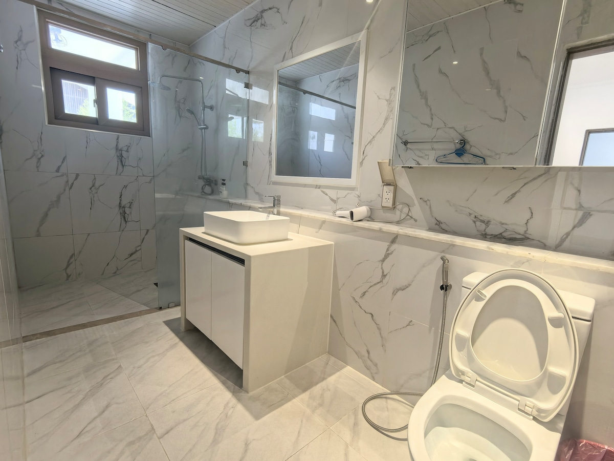 A modern bathroom features marble-patterned tiles on the walls and floor. A large mirror is mounted above a sleek white sink with a stylish faucet. Natural light enters through two windows, and a glass shower area is visible to the left.