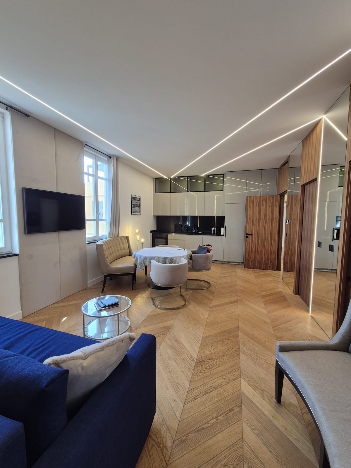 A modern living space features sleek cabinetry and a reflective wall, with a dining table set for two. A blue sofa offers comfortable seating, while a mounted television is positioned for viewing. Natural light filters through large windows, enhancing the contemporary design.