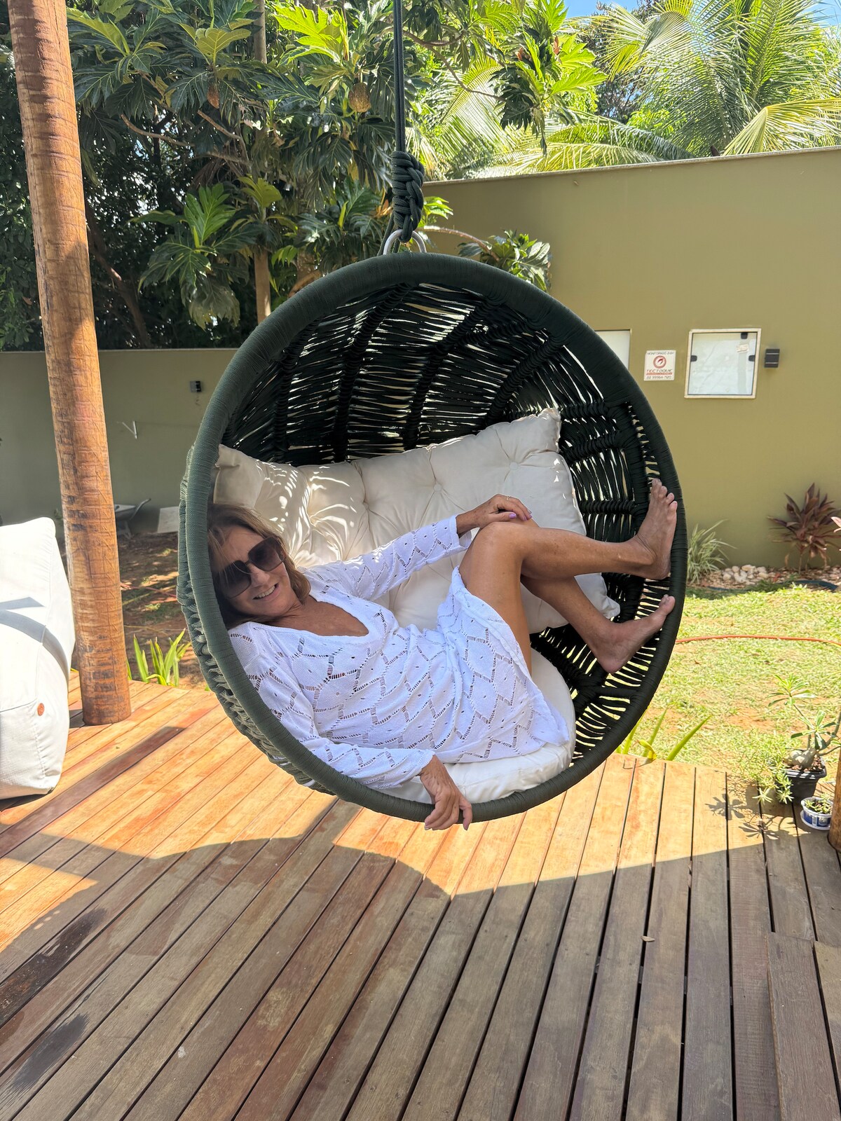 A circular hanging chair is situated on a wooden deck, surrounded by lush greenery. The chair features a soft cushioned interior, inviting relaxation. Sunlight filters through nearby trees, creating a calm atmosphere for outdoor enjoyment.
