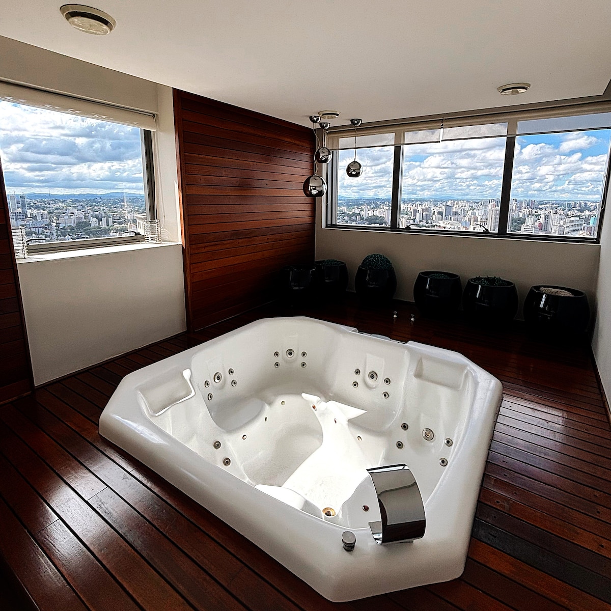 A spacious spa area features a large hot tub surrounded by wooden paneling. Large windows offer panoramic views of the city skyline, allowing natural light to fill the room. Seating arrangements are present along the walls, adding to the relaxation space.