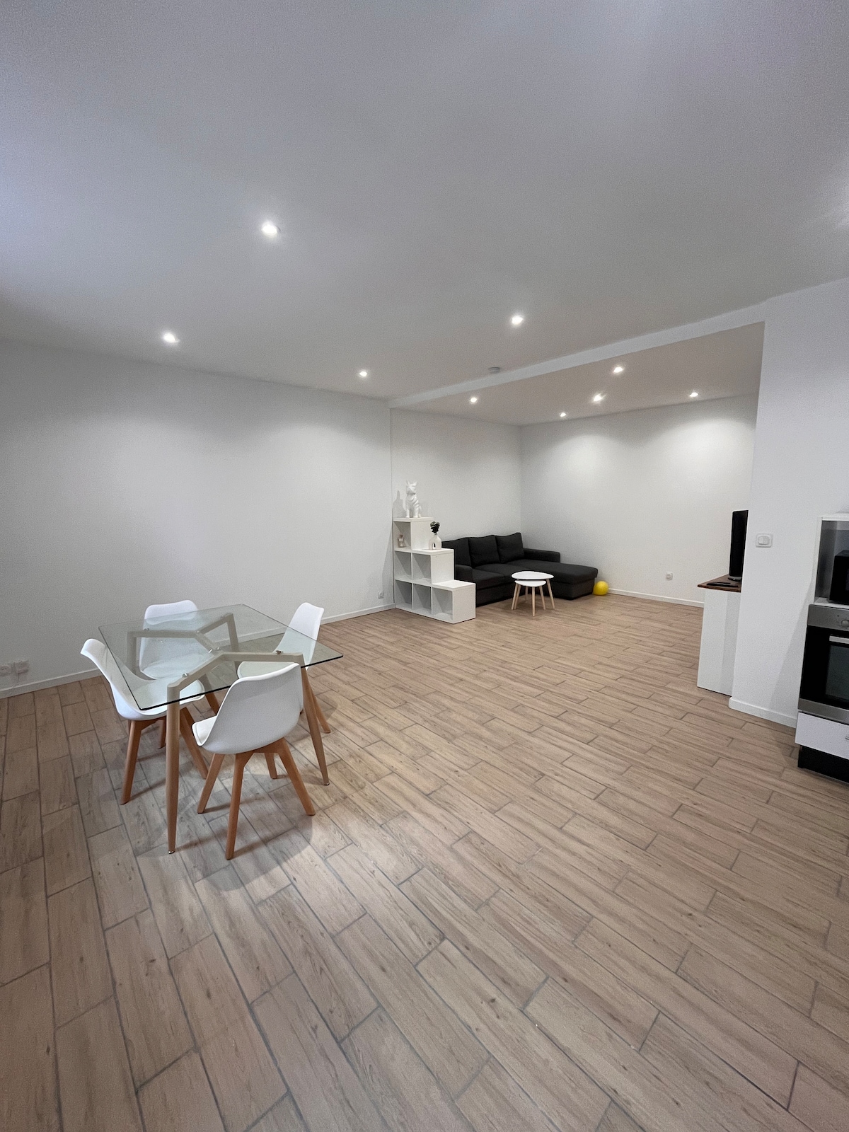 An open living area is presented, featuring a dining table with four white chairs. A black sofa is visible against the wall, complemented by a white shelving unit. Soft lighting from recessed fixtures enhances the spacious feel of the tiled floor.
