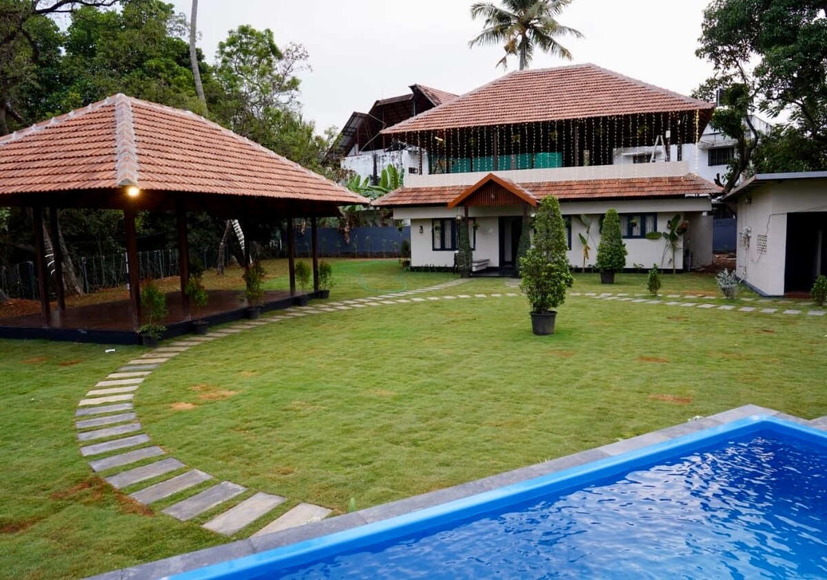 The image shows a well-maintained yard featuring lush green grass bordered by a stone pathway. Two thatched-roof gazebos are visible, along with a blue swimming pool in the foreground. The villa is seen in the background, framed by tall trees.