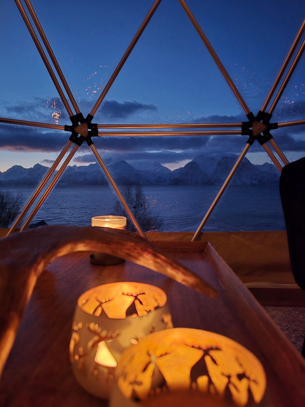 A geodesic dome offers a stunning view of mountains against a twilight sky. Candle holders with intricate designs rest on a wooden surface, softly illuminating the space. The tranquil lake is visible, creating a serene atmosphere as the natural light dims.