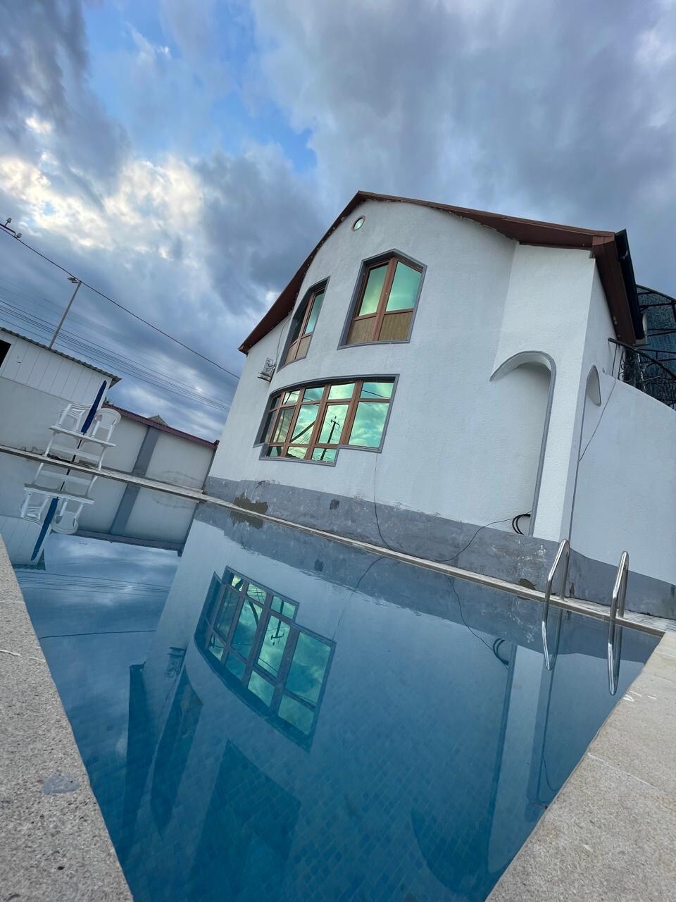 A modern two-story house is positioned beside a clear swimming pool. The structure features large windows reflecting the cloudy sky, while the pool's surface mirrors the architecture. A ladder leads into the pool, enhancing the inviting swimming space.