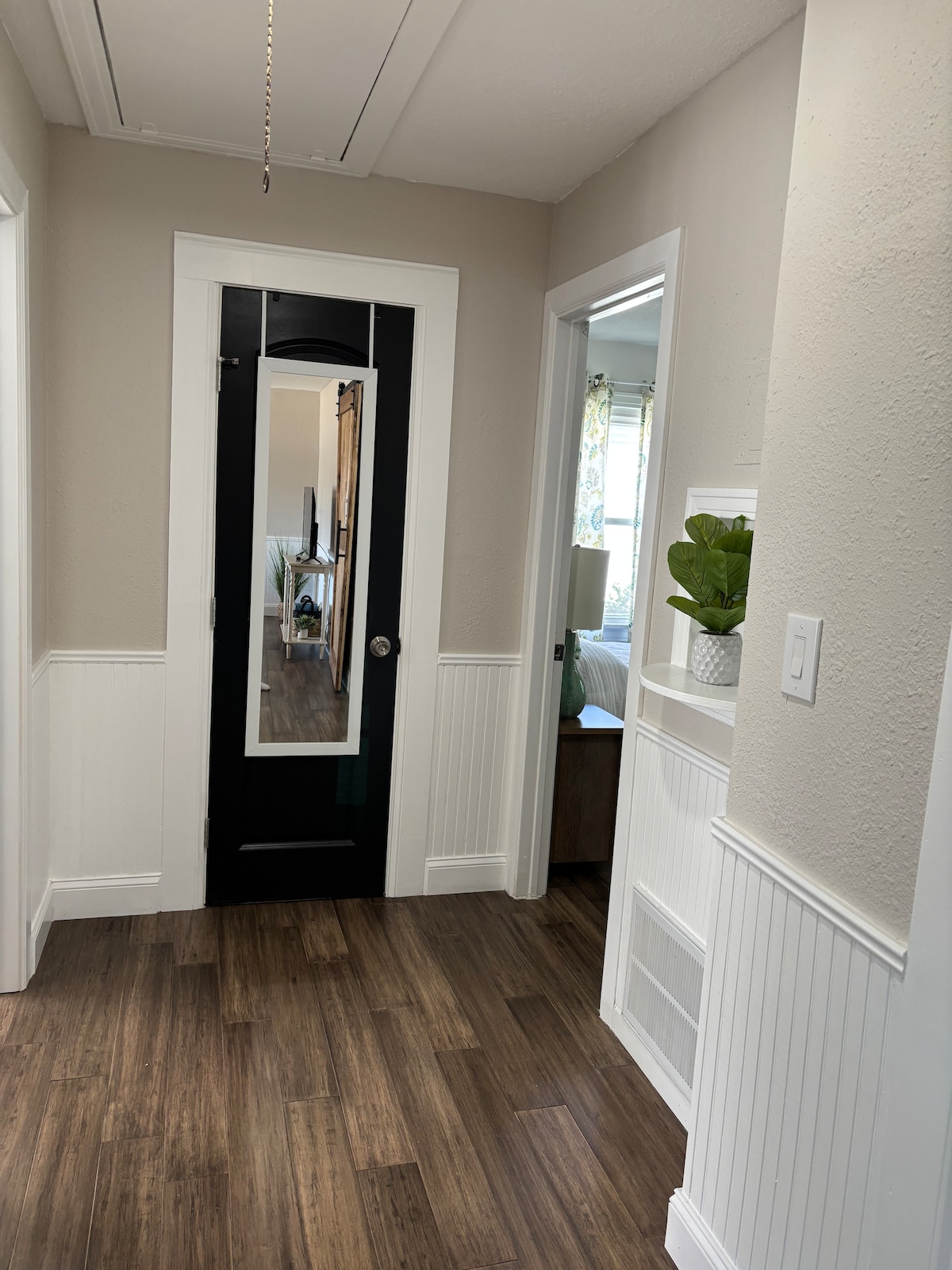 A hallway is depicted, featuring a black door with a mirror that reflects the surroundings. Smooth, wood-like flooring continues throughout. A potted plant is positioned on a shelf against the wall, contributing to the minimalist design. Light enters from the adjacent rooms, enhancing the space.