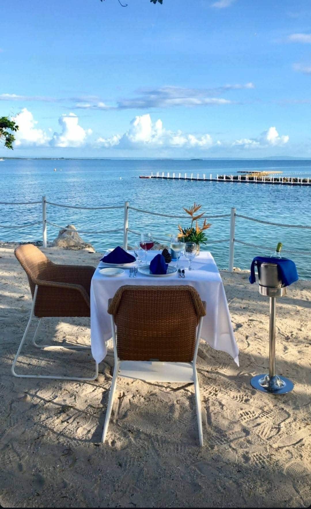 A dining setup is presented on a sandy beach, featuring a table with a white tablecloth. Two wicker chairs are positioned around the table, which is decorated with blue napkins and floral arrangements. The serene ocean view includes a wooden pier and a clear sky with scattered clouds.