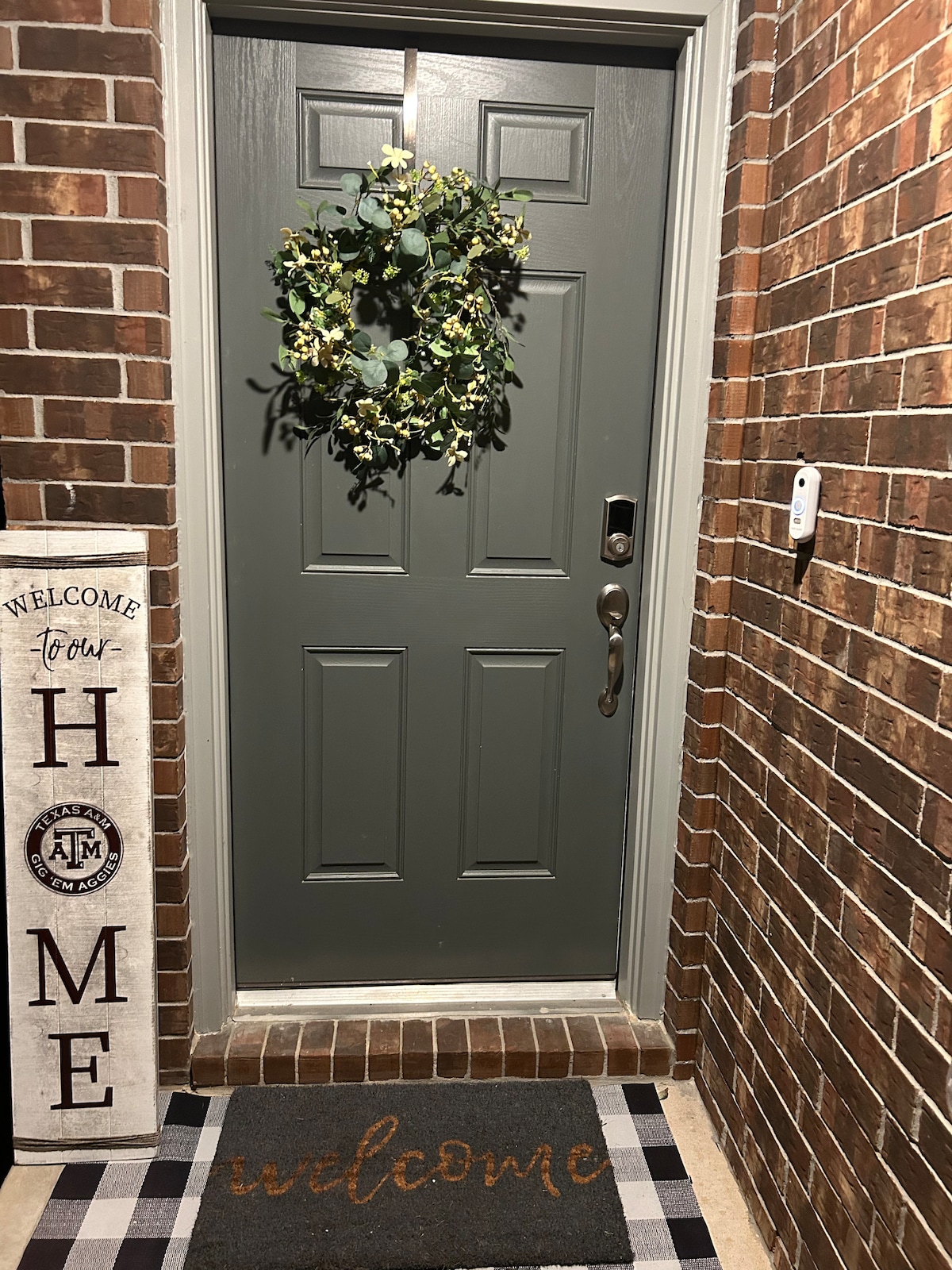 A welcoming entrance is featured with a green front door adorned by a simple wreath. A textured welcome mat sits in front of the door, complemented by a nearby sign that reads 'WELCOME to HOME' against a brick wall.