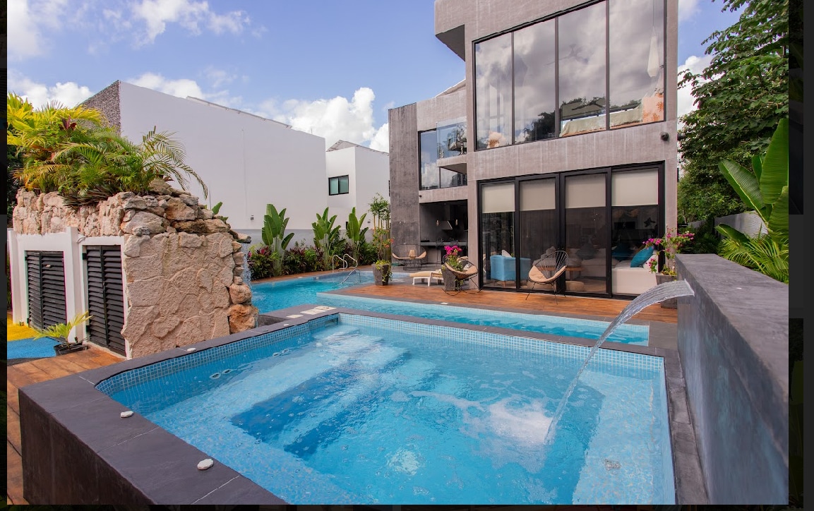 A private pool is surrounded by lush greenery, featuring elegant stone accents. The modern home is showcased with large glass windows, providing a view of the inviting outdoor spaces. Comfortable seating areas are visible alongside the pool, enhancing the tranquil ambiance.