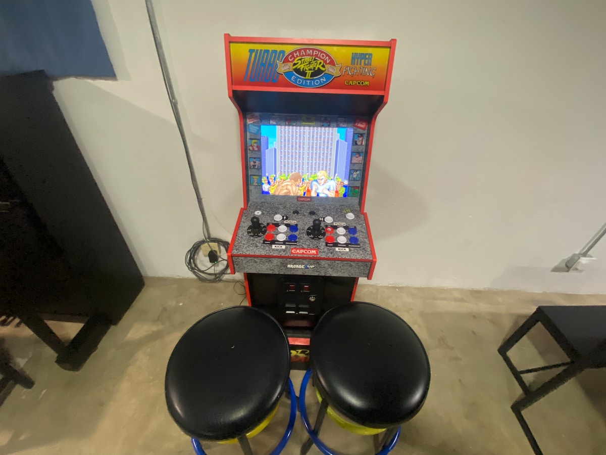 An arcade gaming machine is positioned against a plain wall, featuring a vibrant graphic design. Two black round stools are situated in front of the console, providing seating for players. The machine displays colorful gameplay visuals, inviting interaction.