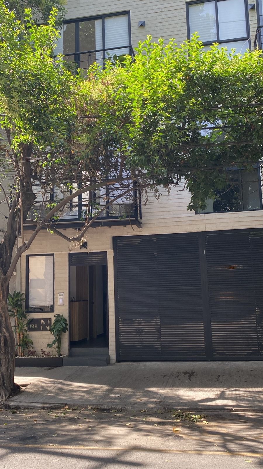 An urban building is shown with a modern design featuring large windows and a covered entrance. Lush greenery frames the facade, enhancing the natural ambiance. A solid gate and subtle architectural details contribute to the overall contemporary look of the structure.