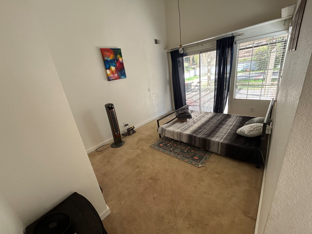A spacious room features a low bed with a striped blanket and a decorative rug on the floor. Large windows provide natural light and a view of the landscaped backyard. A painting in vibrant colors decorates the wall, adding a touch of creativity to the environment.