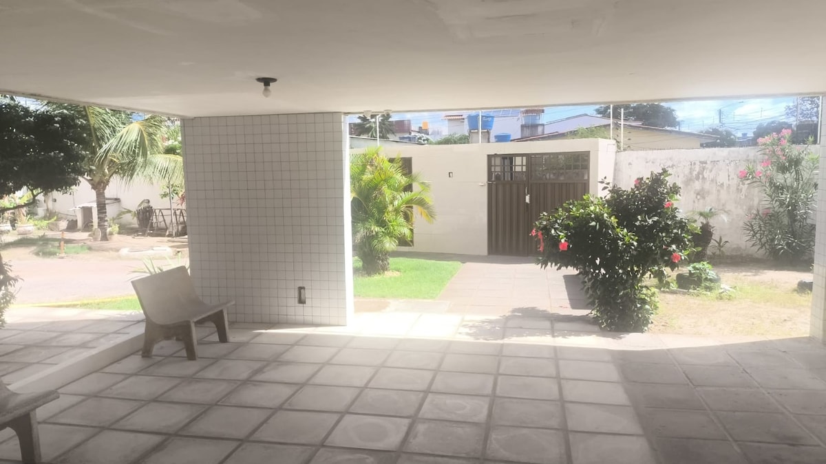 The entrance area is designed with large tiles and features a covered space with a bench. Greenery, including flowering plants, enhances the surroundings, while a gate leads to the garden area beyond. Natural light brightens the space, creating an open and welcoming feel.
