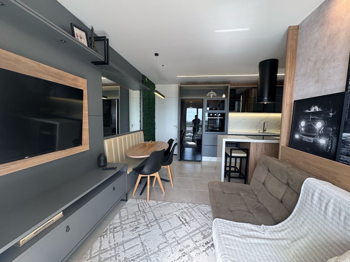 A modern living space showcases a sleek television unit with wooden accents, complemented by a circular dining table surrounded by four chairs. A comfortable sofa is placed next to a textured area rug, while the kitchen area is visible in the background, featuring dark cabinetry and appliances.