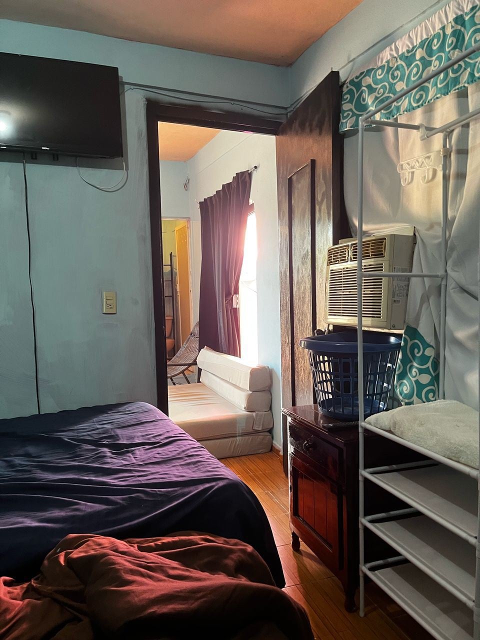 A simple bedroom is depicted, featuring a spacious bed with a dark blanket. A wall-mounted television is visible above, and an air conditioning unit is mounted in the window. A doorway leads to another room, while a laundry basket sits on a nearby shelving unit.