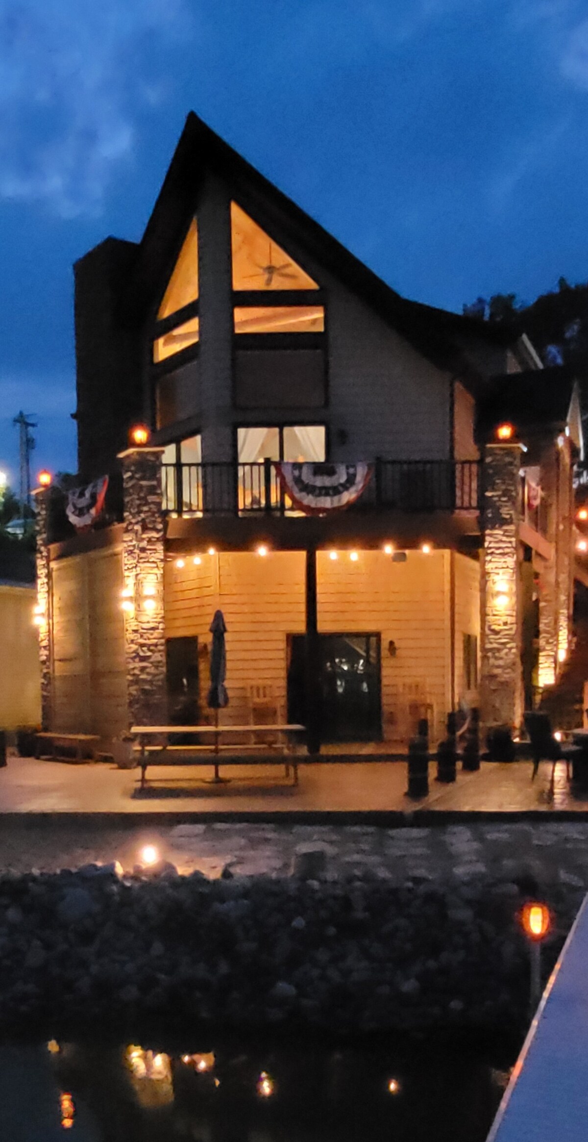 An elegant lakeside home is showcased at twilight, featuring a stunning A-frame design with large front windows. Soft lighting illuminates the exterior, highlighting the stone and wood accents. Festive decorations adorn the balconies, and a spacious patio area is visible near the water.