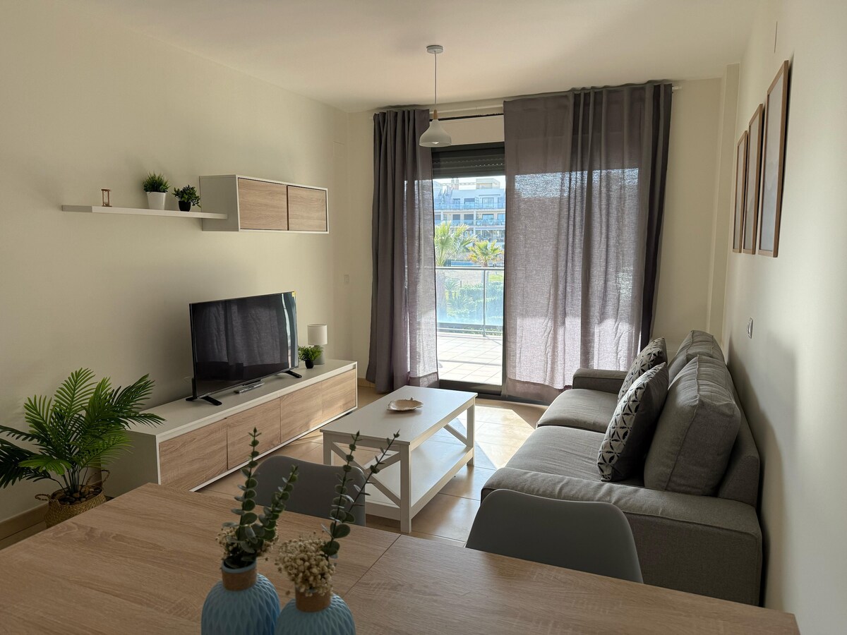 A bright living room features a comfortable gray sofa positioned beside a wooden coffee table. A television is mounted on the wall opposite the sofa, with a decorative shelf above it. Large glass doors lead to a balcony, letting in ample natural light.