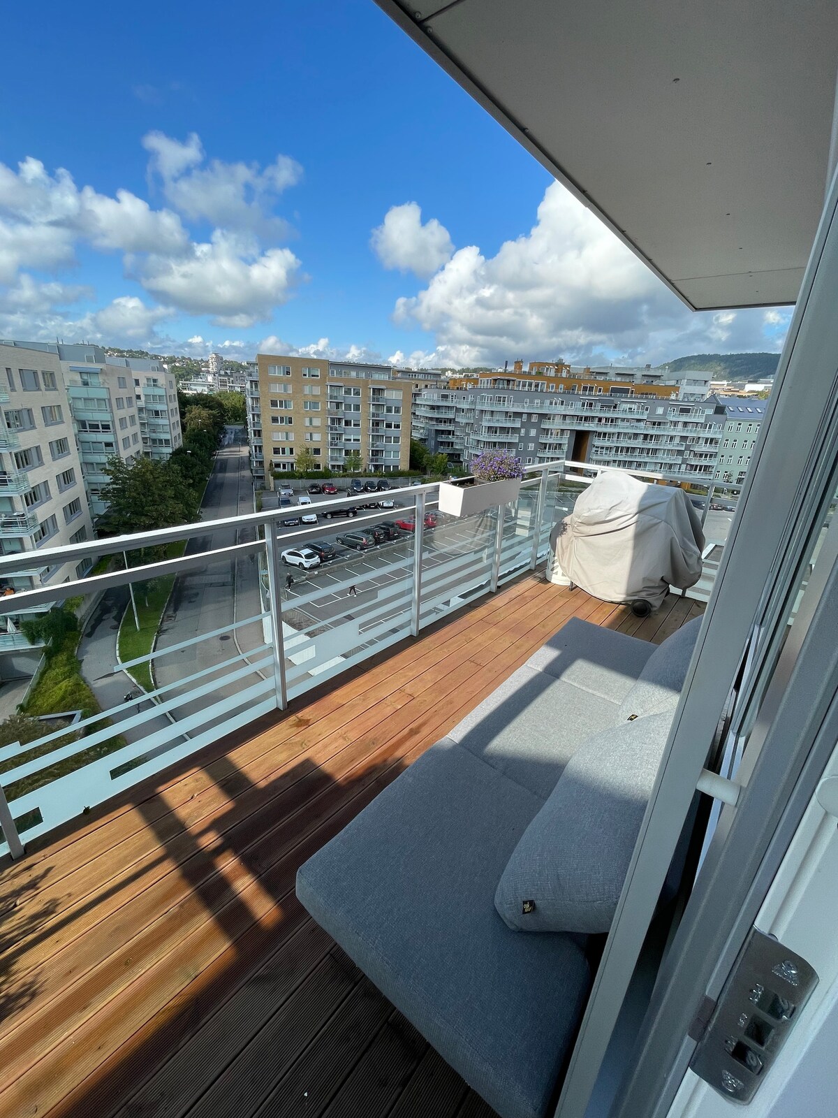 A spacious balcony is featured with wooden decking and a comfortable outdoor sofa. A view overlooks the street and nearby buildings, with blue skies and scattered clouds above. The balcony provides ample space for relaxation while enjoying the surroundings.