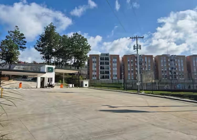 A spacious open area is captured, featuring a well-maintained entrance to an apartment complex. Several buildings with large windows and balconies are visible in the background, framed by trees and a clear blue sky, while a roadway is seen leading into the property.