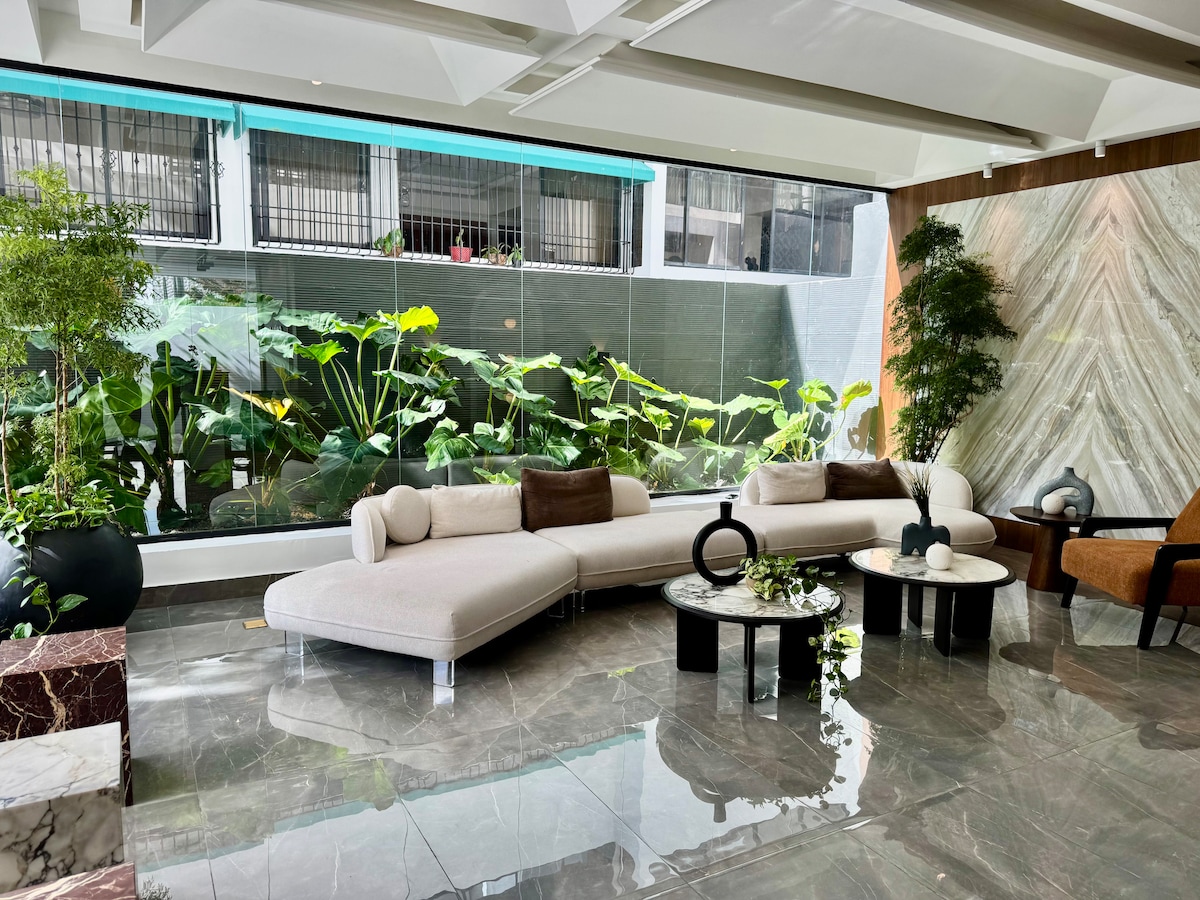 A spacious lounge area features a curved sectional sofa in soft tones, complemented by a marble coffee table. Floor-to-ceiling windows provide a view of lush green plants, enhancing the natural light that fills the space. Decorative elements include minimalist vases and a comfortable armchair.