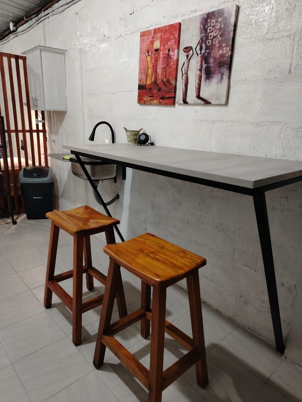 A minimalist workspace is depicted against a white brick wall, featuring a concrete tabletop supported by black metal legs. Two wooden stools provide seating, and two framed artworks add color to the wall above the table. A small shelf and waste bin are visible in the background.