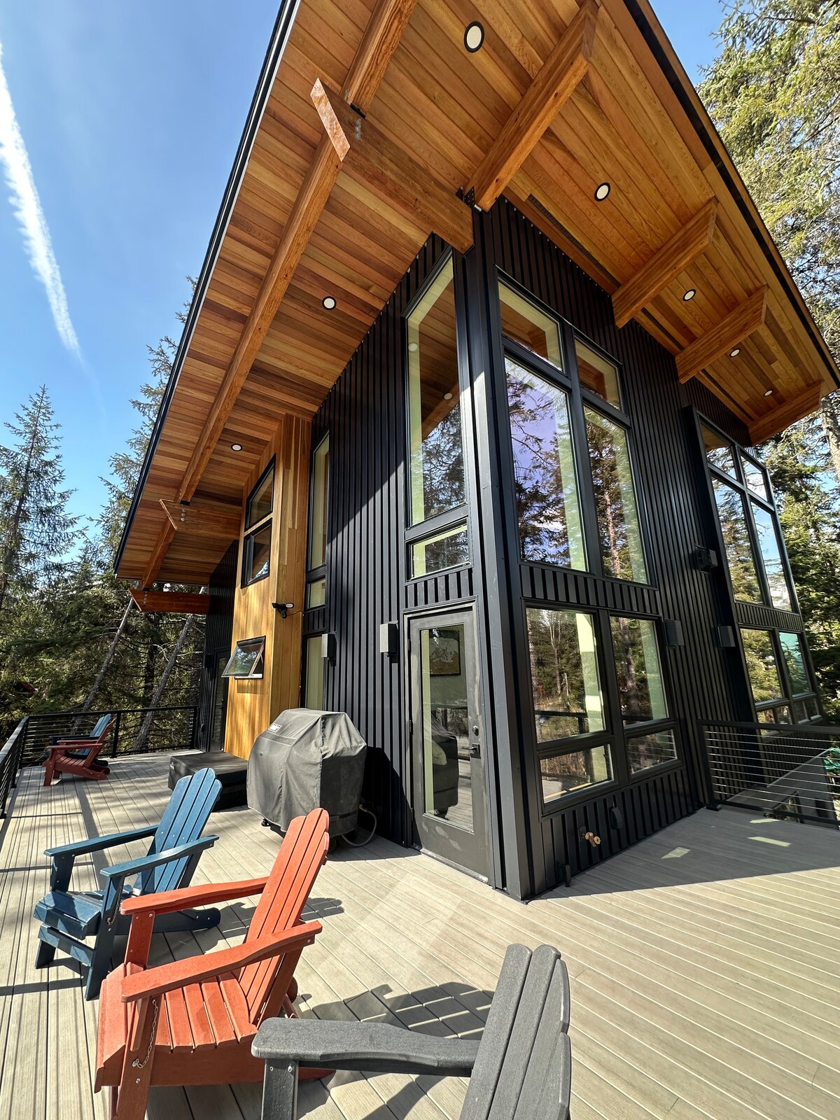 The exterior of the mountain modern chalet is showcased, featuring a combination of wood and black metal siding. Large windows enhance natural lighting. A spacious deck is positioned in front, furnished with colorful outdoor chairs and a gas grill for outdoor dining.