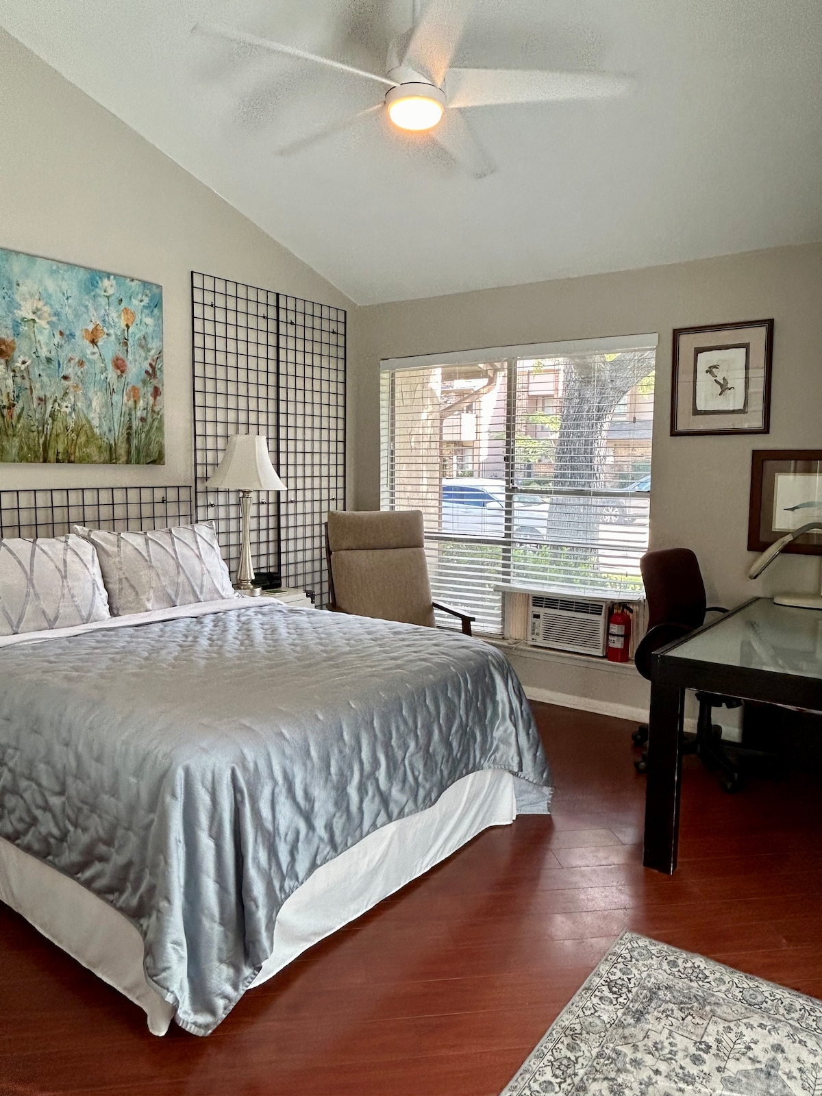A tranquil sleeping area features a spacious bed with a gray quilt, complemented by light-colored pillows. A soft armchair and a desk are positioned by the window, which allows natural light to illuminate the room. Decorative wall art adds a touch of elegance.