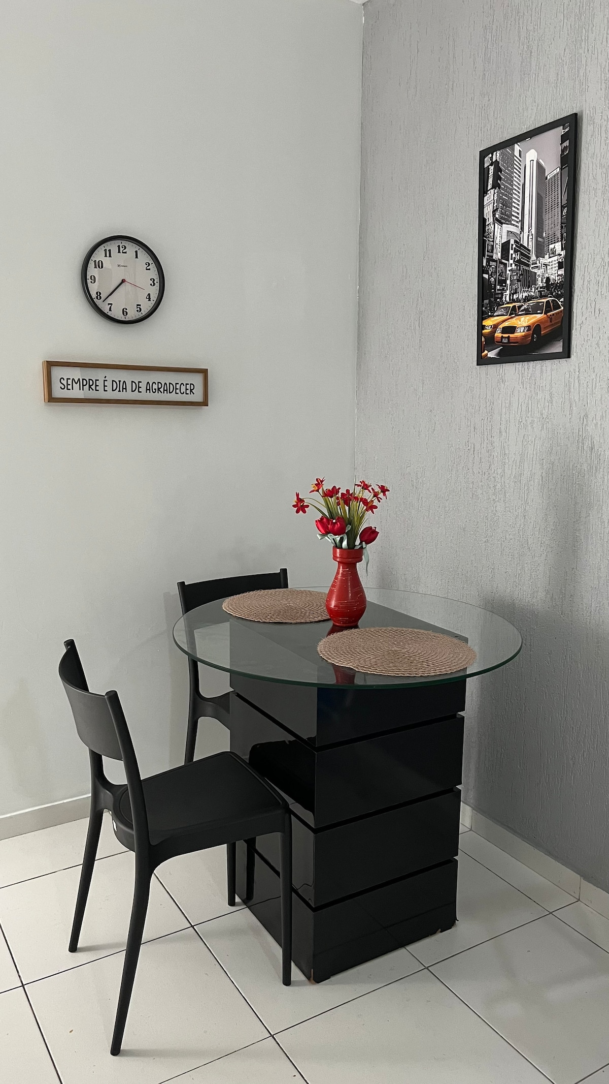 A small dining area is presented with a round glass-top table supported by a black base. Two black chairs are positioned beside the table. A vase of flowers adds a pop of color to the setting, and a wall clock and framed photo provide visual interest.