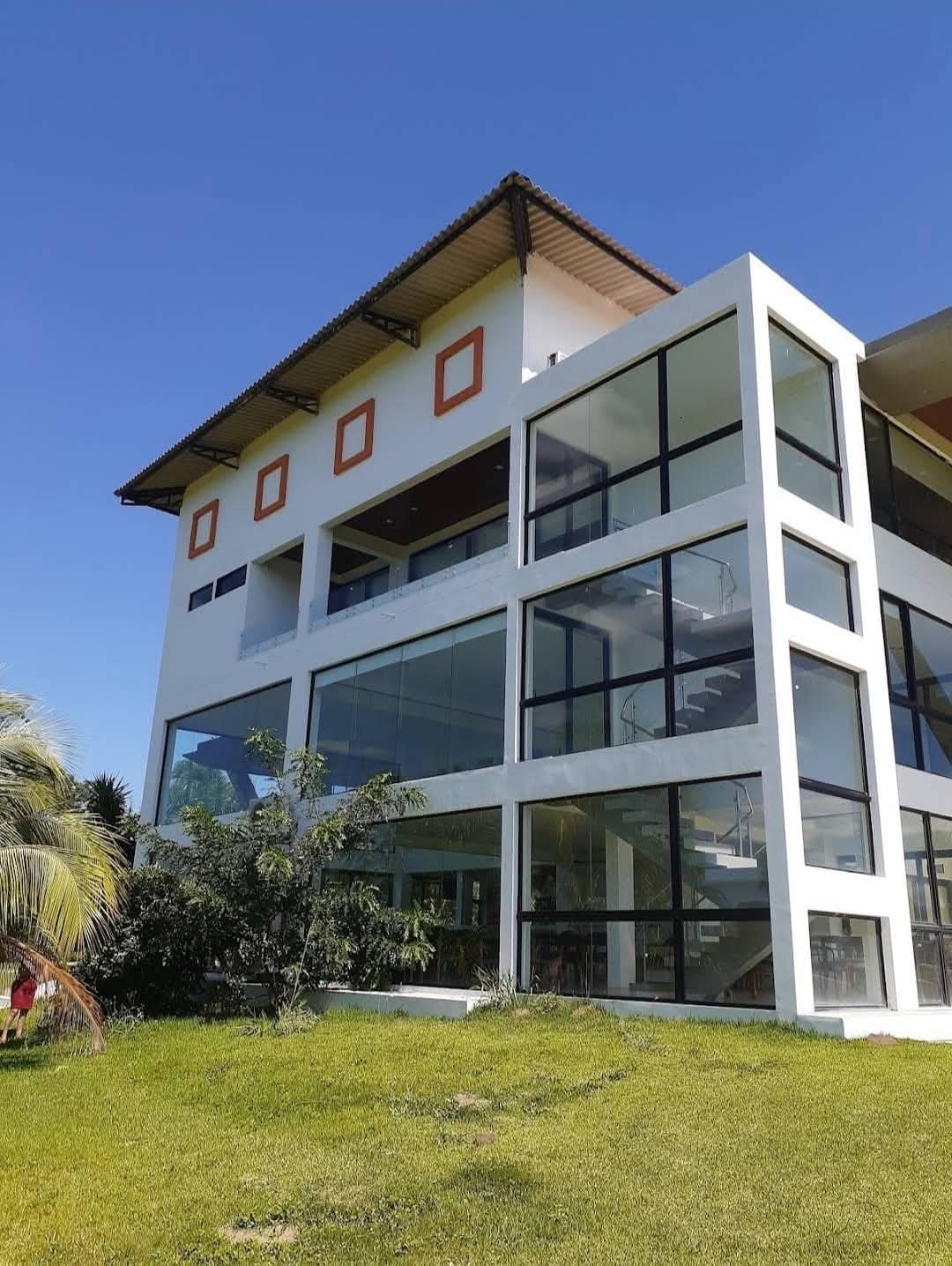 A modern three-story building is showcased, featuring large glass windows that allow for ample natural light. A lush green lawn surrounds the base, and a staircase is visible through the transparent facade. The sky is clear and blue, adding to the serene setting.