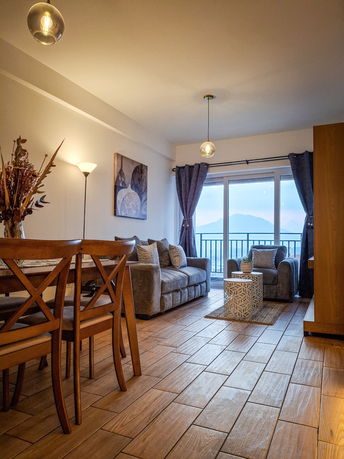 An open living space features a comfortable sofa and a sofa bed, complemented by a small table. Natural light enters through a large window, offering a view of the mountains. A dining area with wooden chairs is visible, creating a warm and inviting atmosphere.