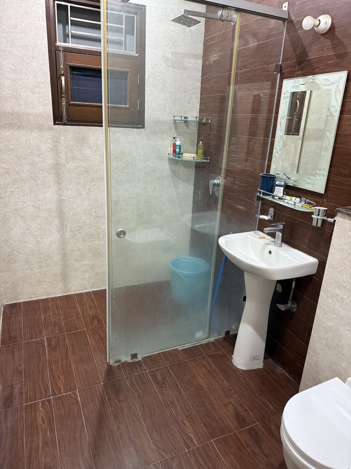 A bathroom is presented with a glass shower enclosure and a modern sink. A mirror is mounted above the sink, and basic toiletries are arranged on a shelf. Natural light enters through a small window, while the flooring features a rich brown tone.