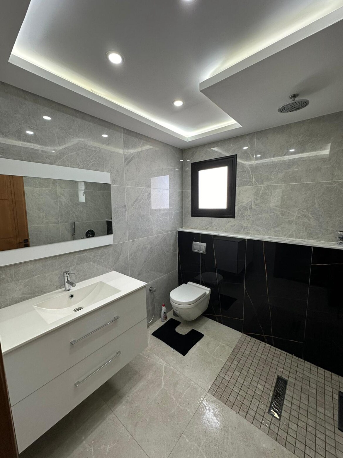 A modern bathroom features elegant gray stone walls and a stylish black accent wall. A white vanity with dual drawers is paired with a mirror above. Bright lighting illuminates the space, and a compact toilet is positioned beside a small window.