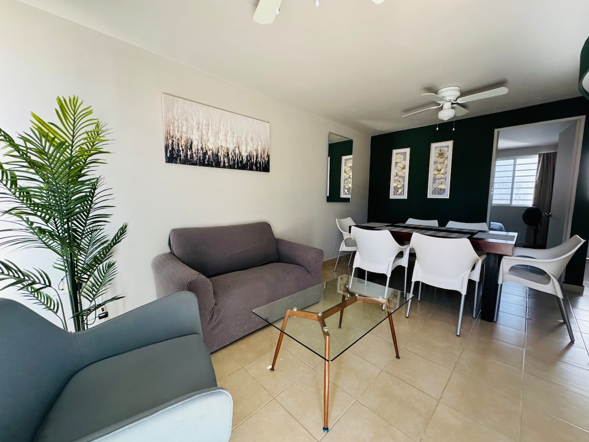 A modern living space is shown with a gray sofa and an additional light blue chair. A glass coffee table rests on the tiled floor, surrounded by a wooden dining table and white chairs. Wall art and indoor plants add subtle decor to the area.