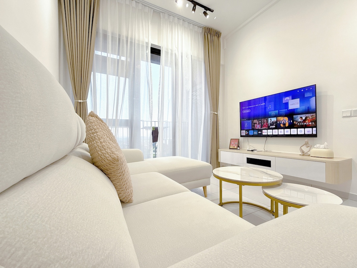A light-filled living area features a plush beige sectional sofa, accentuated by a textured pillow. A sleek television is mounted on the wall, displaying vibrant content. Two modern coffee tables are positioned in front, and sheer curtains allow natural light to softly illuminate the space.