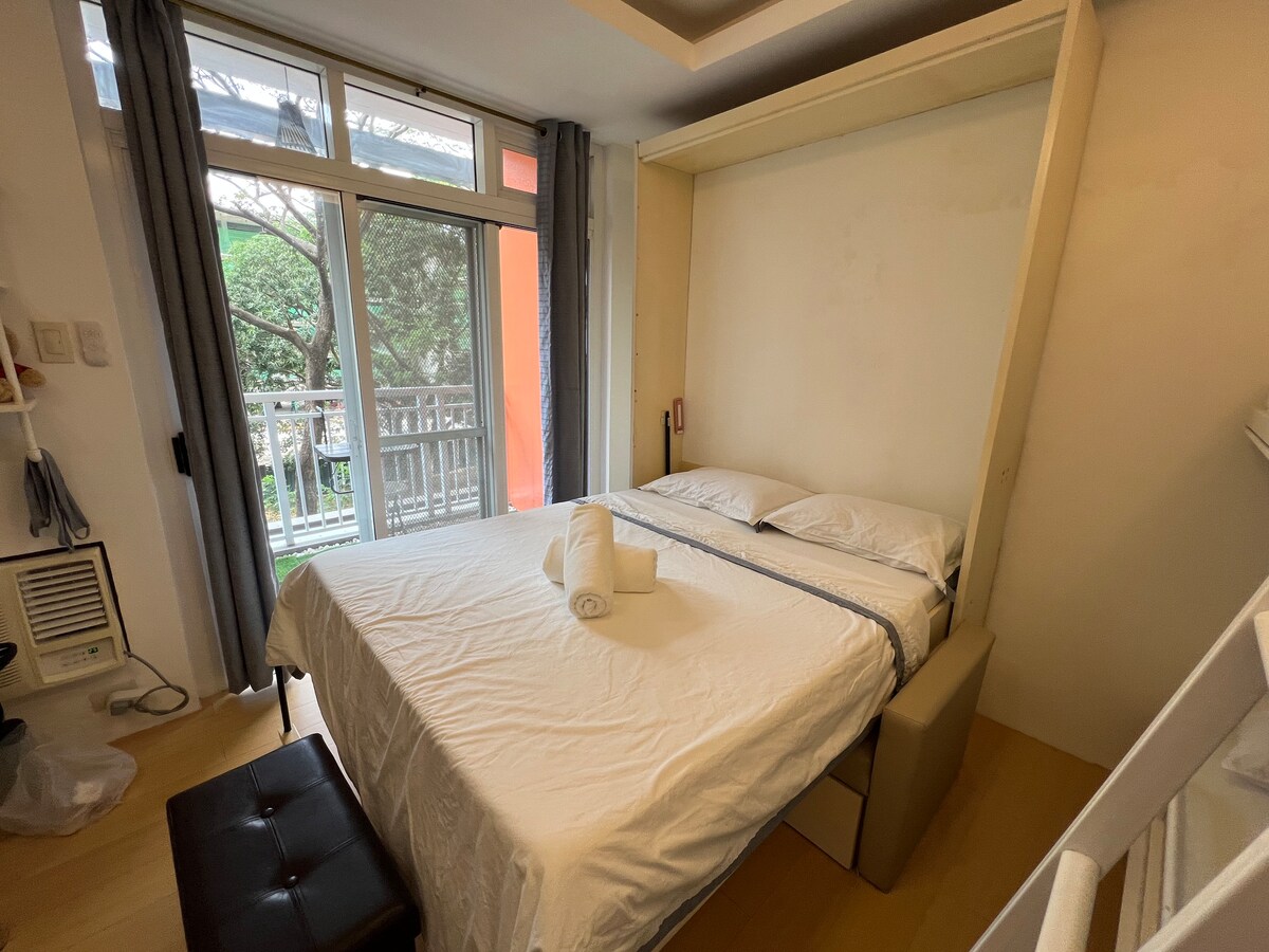 A compact bedroom features a spacious wall bed that folds up to optimize space. Natural light filters in through large windows adorned with curtains, offering views of greenery outside. A neatly arranged towel is placed on the bed, and a small ottoman is positioned at the foot.