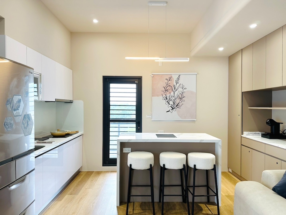 The kitchen features a modern design with sleek cabinetry and a spacious island. Three bar stools are positioned at the island, complemented by a contemporary pendant light above. A window provides natural light, enhancing the overall bright feel of the space.