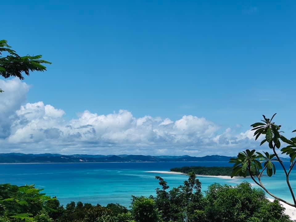 A panoramic view of a sandy beach transitions into vibrant turquoise waters, framed by lush greenery. The horizon is defined by gentle waves and a clear blue sky adorned with scattered clouds, enhancing the serene coastal landscape.