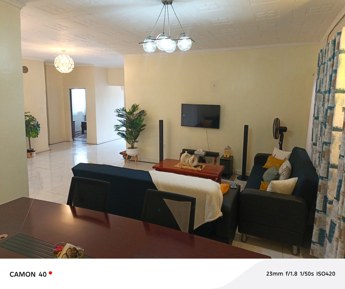 A spacious living area is presented, featuring a dark sectional sofa and a wooden coffee table. A television is mounted on the wall. Nearby, a green plant adds a touch of nature, while light-colored curtains frame a large window, enhancing the overall brightness.
